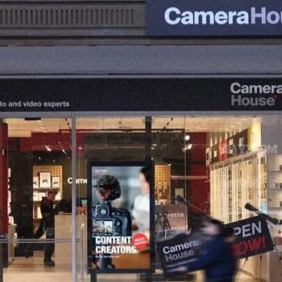 Storefront of a Camera House retail location with large glass windows, featuring prominent Camera House signage and digital displays, and a blurred pedestrian walking past.