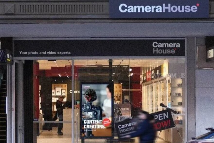 Storefront of a Camera House retail location with large glass windows, featuring prominent Camera House signage and digital displays, and a blurred pedestrian walking past.