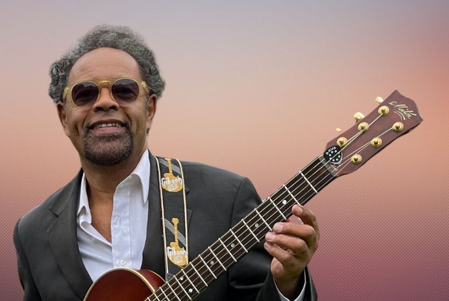 Singer and guitarist Little Paul, with sunglasses on, guitar in hand, smiling. 