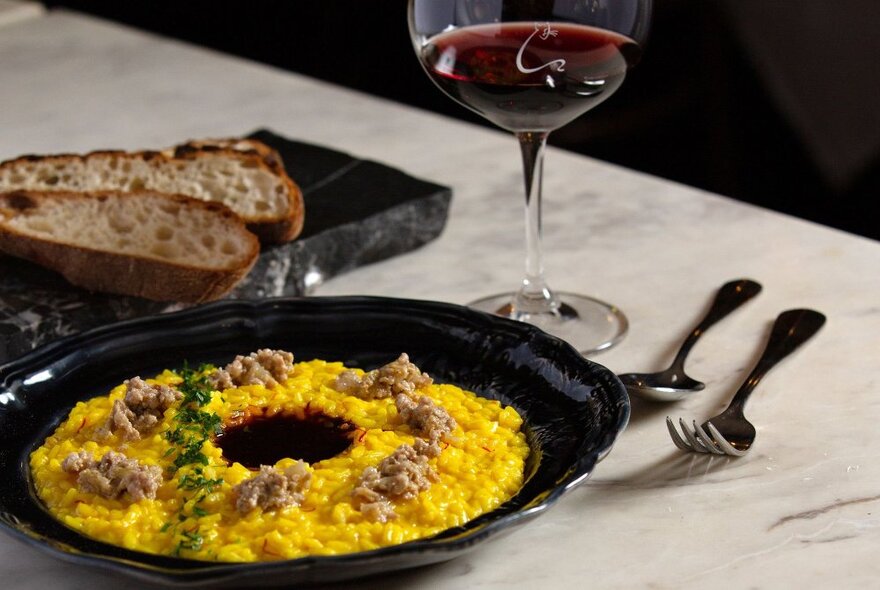 A dish of saffron infused risotto, a glass of red wine and two slices of bread on a marble topped table.