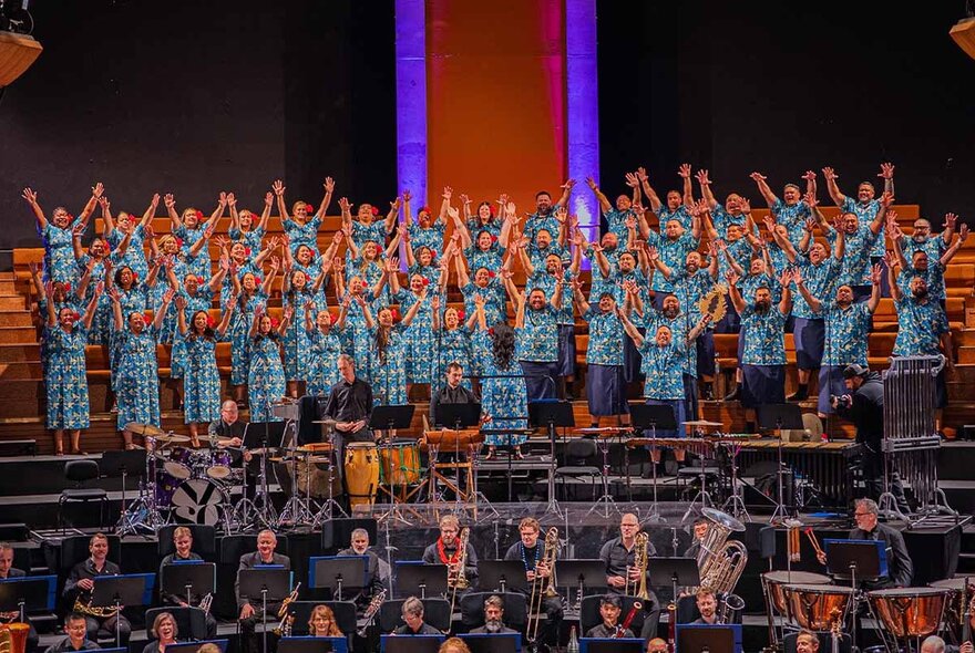 A Pacific nations choir on stage all wearing blue with their hands raised in the air, an orchestra in the foreground. 