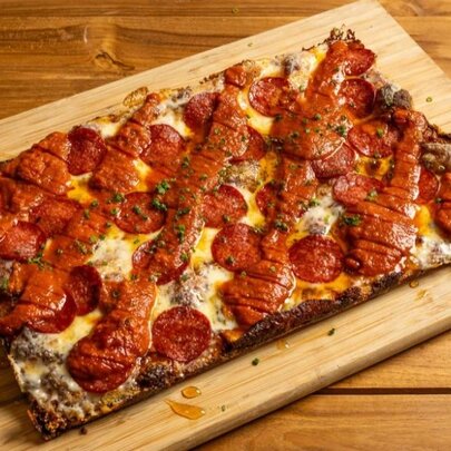 A rectangle pizza with pepperoni and a chilli sauce drizzed over it, presented on a rectangle wooden board.