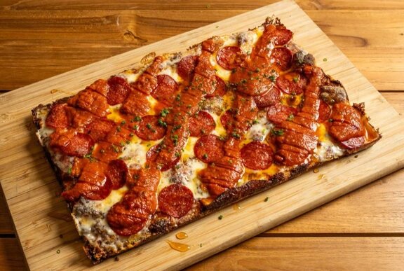 A rectangle pizza with pepperoni and a chilli sauce drizzed over it, presented on a rectangle wooden board.