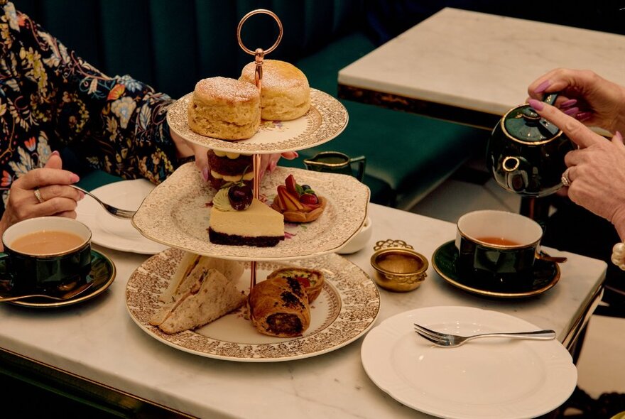People seated at a cafe table enjoying a high tea, with a hand pouring tea from a pot into a cup, and sweet and savoury treats arranged on a three-tiered cake stand.
