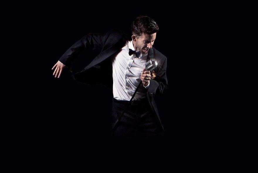 Man wearing a tuxedo singing into a microphone with his right arm swung out behind him, against a black background.