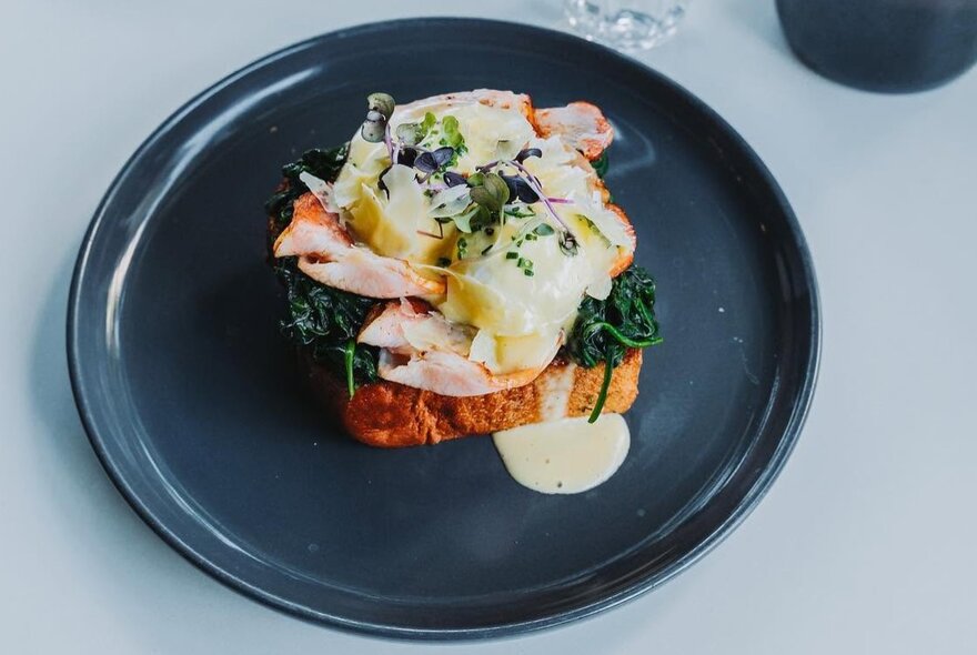 Eggs Benedict-style brunch plate featuring a poached egg, smoked salmon, and hollandaise sauce served over toast, on a dark plate.