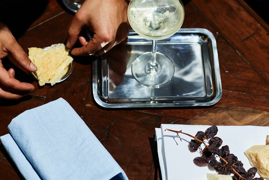 Hands picking up cheese and crackers from a small plate, a martini on the table nearby, plus a cheese plate with dried grapes.