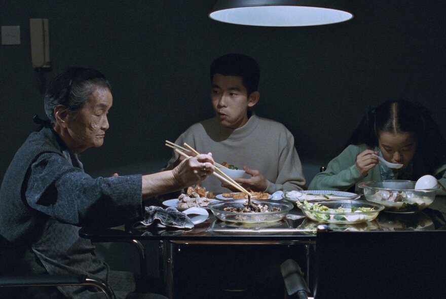 Still from the film Autumn Moon, with three people seated at a table eating food in a dimly lit room.