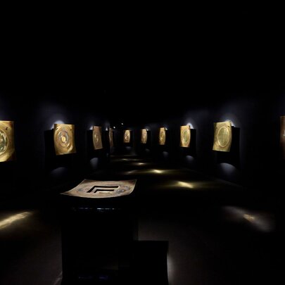 Large brass percussion instruments installed on a wall in a darkened gallery space. 