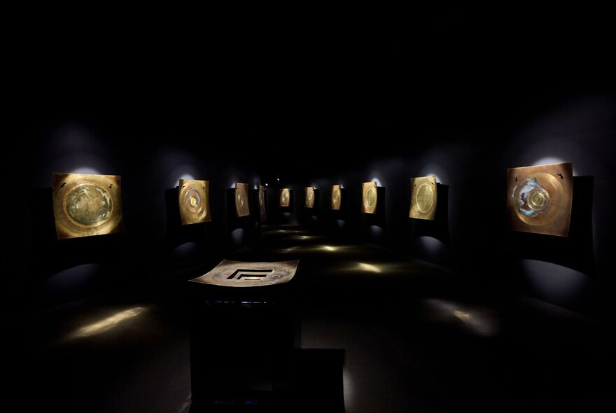 Large brass percussion instruments installed on a wall in a darkened gallery space. 