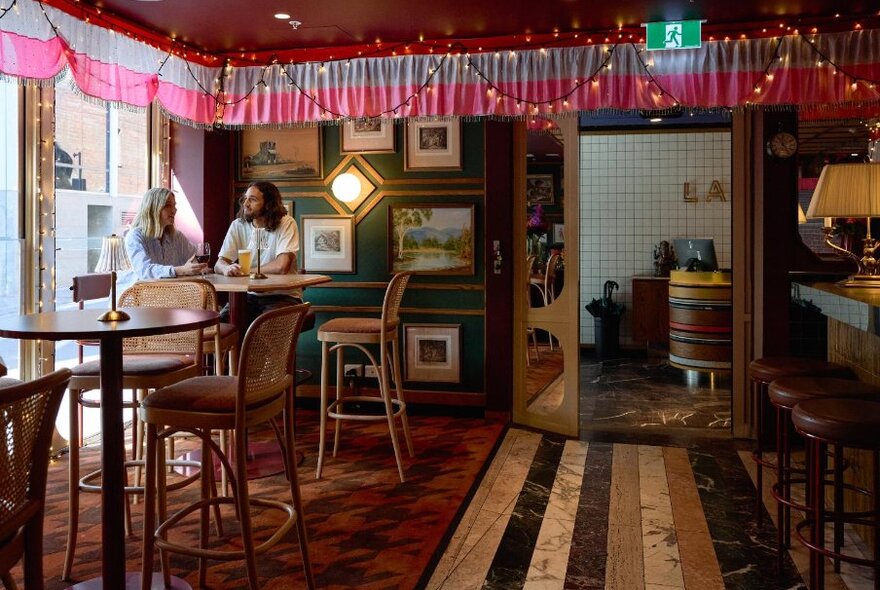 Two people in a highly decorated pub dining room with fairy lights and pink curtains.