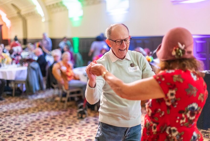 An older couple dancing together in a banquet hall with rainbow coloured lights behind them.