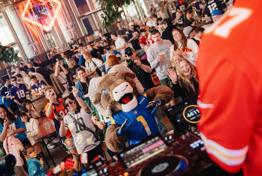 A crowded indoor venue filled with football fans, featuring the Los Angeles Rams mascot, Rampage, wearing a blue jersey and operating a DJ setup, while a person in a bright orange or red jersey stands in the immediate right foreground facing the crowd.