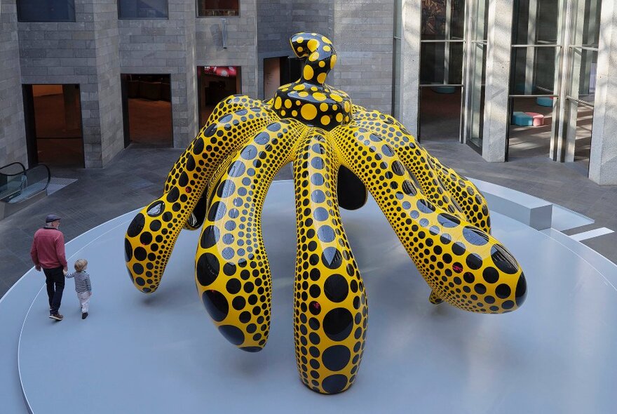 A parent and child walking past a giant abstract pumpkin sculpture painted yellow with black spots.