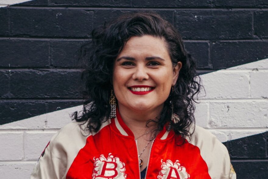 Dr Paola Balla is a Wemba Wemba and Gunditjmara woman, wearing a cream and red initialled bomber jacket.