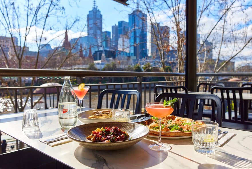 A restaurant outdoors with a table of pizza, pasta and cocktails on the table.