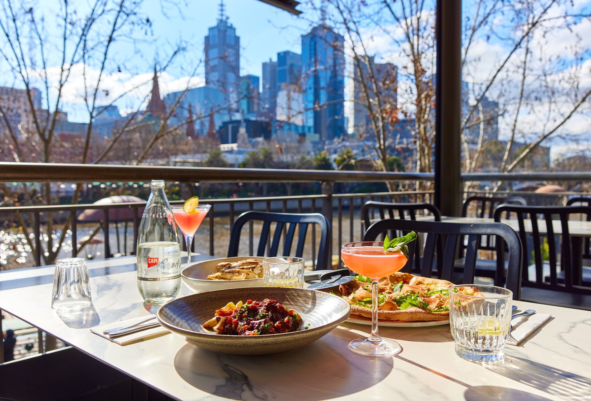 A restaurant outdoors with a table of pizza, pasta and cocktails on the table.