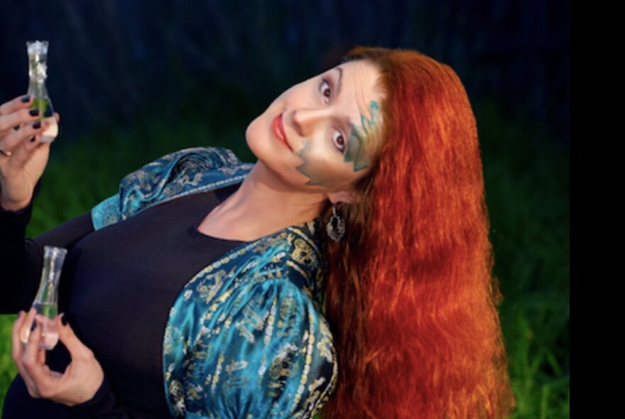 Performer with long red hair, leaning sideways holding two glass bottles, with blue make-up on her face.