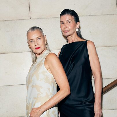 Two middle-aged fashion models wearing sophisticated sleeveless midi dresses, including a cream-colored patterned style and a sleek black satin design, against a white wall.