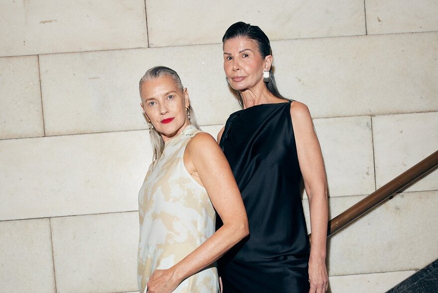 Two middle-aged fashion models wearing sophisticated sleeveless midi dresses, including a cream-colored patterned style and a sleek black satin design, against a white wall.