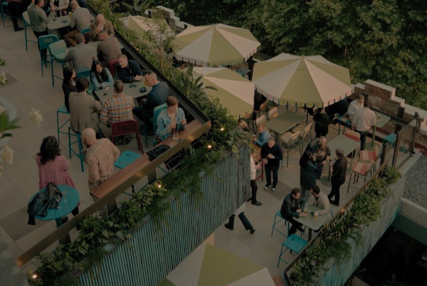 Looking down from above at the two open-air rooftop terraces at The Waterside Hotel, with shade umbrellas, high bar tables, stools and greenery, and people socialising in the spaces.