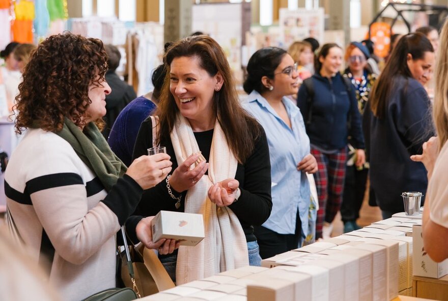 People smiling and looking at items at an indoor design market stall, with crowds of people wandering around in the background.