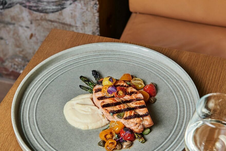 A plate with a pan fried salmon fillet on a salad with a cream coloured sauce, glass of wine in the foreground.