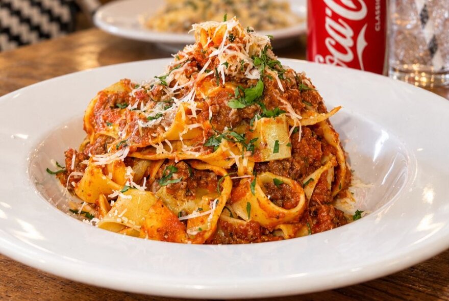 A plated, serving of pasta bolognese topped with parmesan cheese and fresh herbs, sitting on a restaurant table.