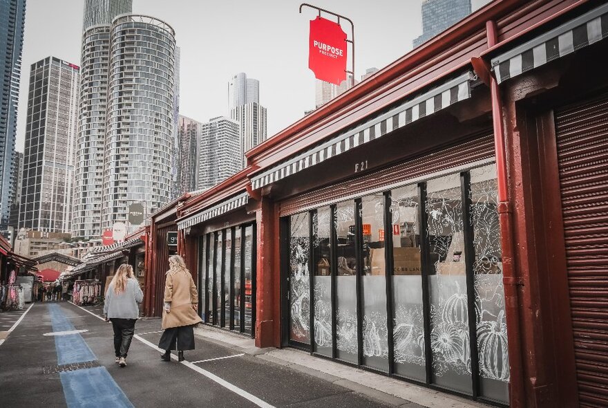 The exterior of the Purpose Precinct stall at Queen Victoria Market, with two people walking by, and city skyscrapers in the background.