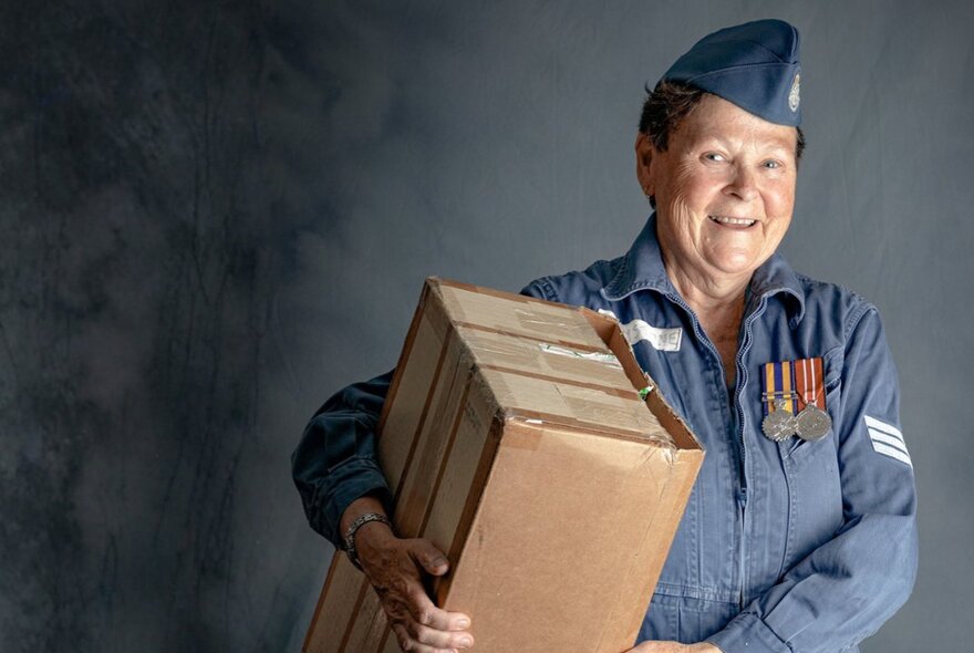 A smiling woman in an Australian military uniform, adorned with medals, holding a large cardboard box.