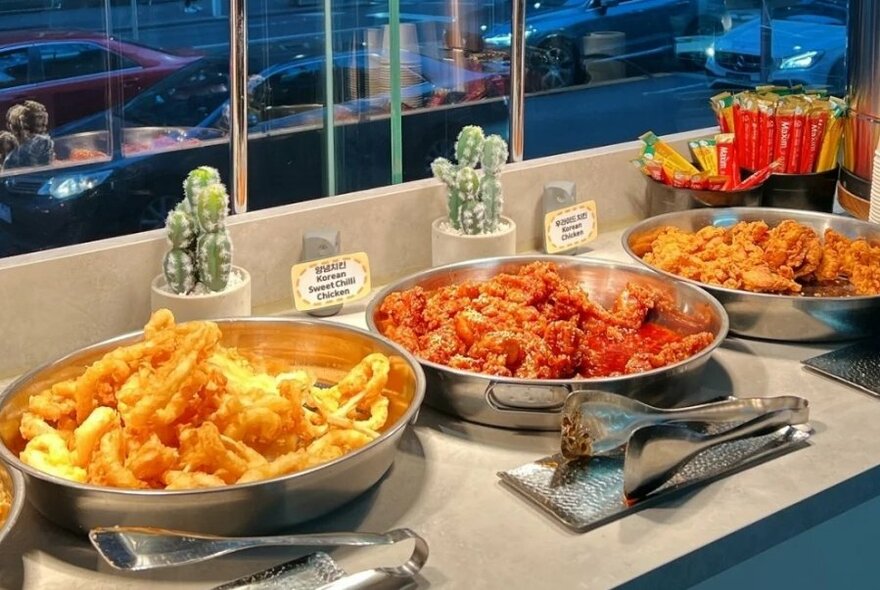 Large round dishes of food on a buffet table with serving tongs in front of them.