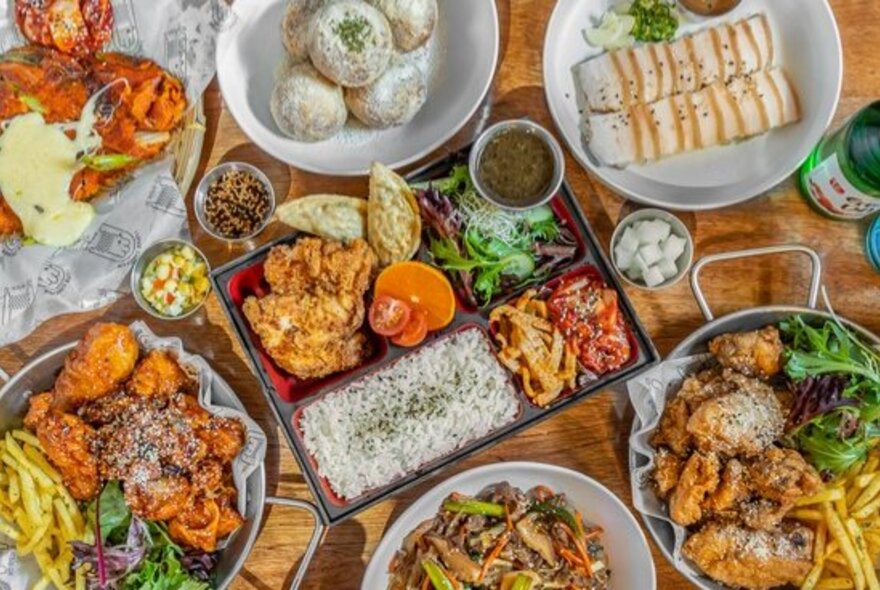 Looking down on a table laden with many plates of Korean chicken and bento boxes. 