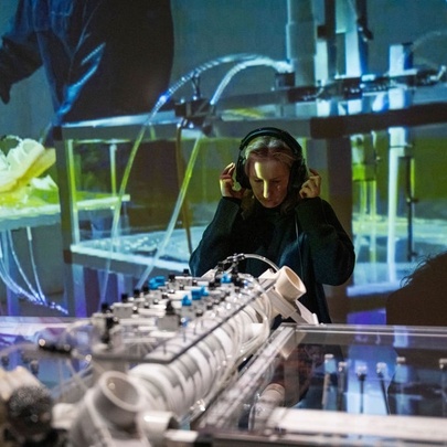 A person with headphones on their head, standing in front of a large instrument, a wall behind them displays an image of hands at a control panel.