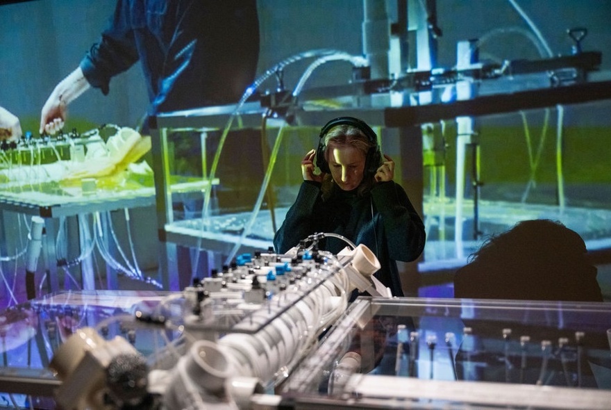 A person with headphones on their head, standing in front of a large instrument, a wall behind them displays an image of hands at a control panel.