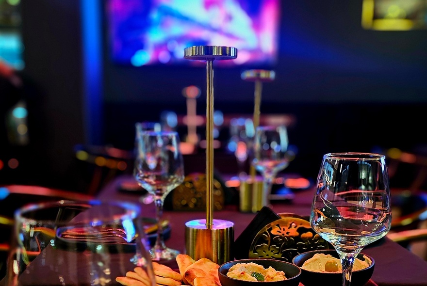Looking down a long table with wine glasses and appetisers, a video screen out of focus in the background. 