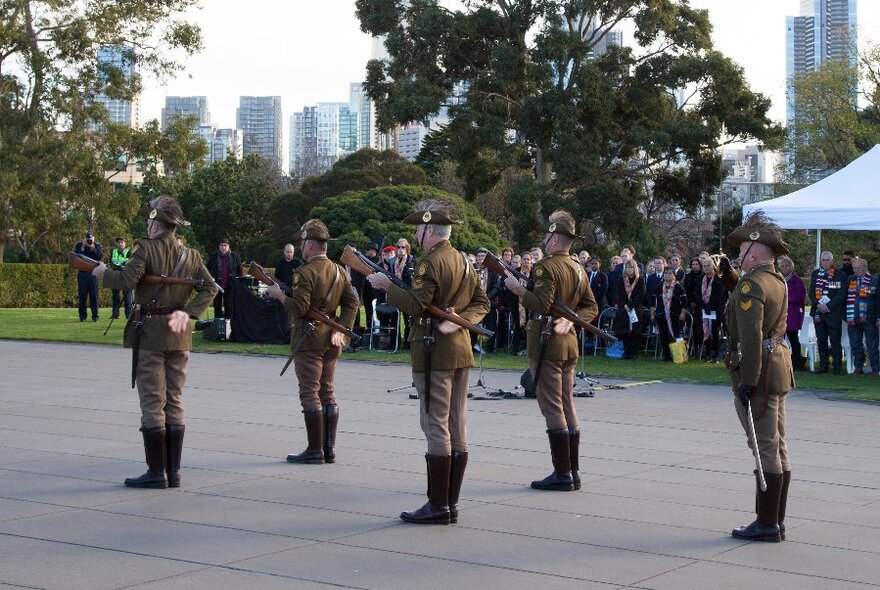 Five soldiers in army uniform stand still in a public area with the city in the background. 