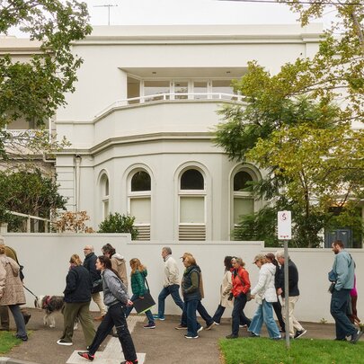 A group of people participating in a guide-led tour, walking past a large white residence and trees, on a local street.