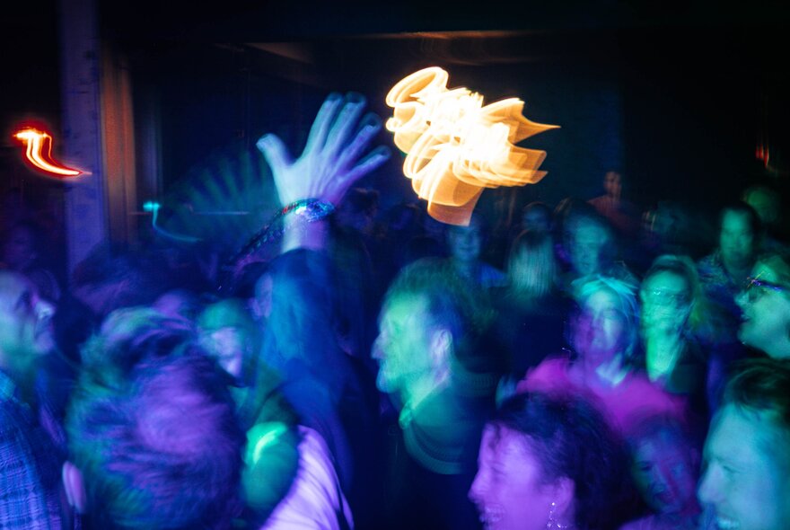 A blurred picture of people on a dancefloor with green and purple lights and a blurred flare of yellow in the middle. 