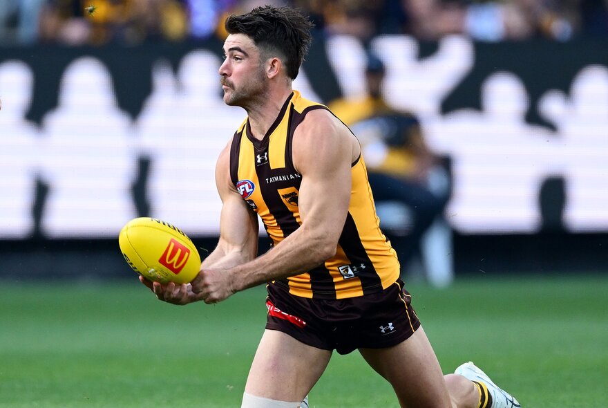 A Hawthorn AFL player running on the field while about to handball a yellow football.