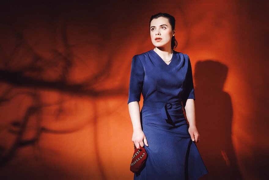 An woman wearing a 1940s style blue dress, with elbow length sleeves, her hair styled away from her face, standing against a dark orange background with the shadow of a tree on it.