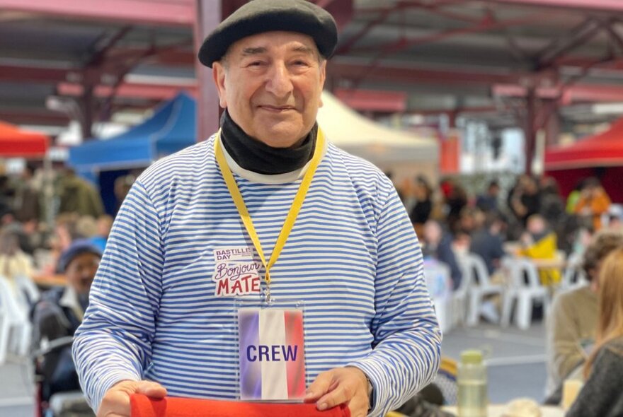 A older men wearing a striped navy and white nautical top with long sleeves, and a black beret, standing in the sheds of Queen Victoria Market.
