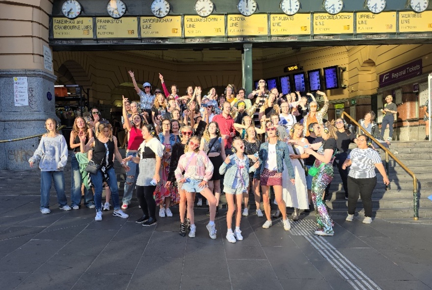 Crowd gathered under Flinders Street Station Clocks and steps, waving arms and pulling poses.