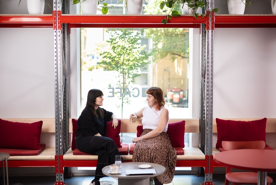 Two people seated and talking on red armchairs at the Science Gallery Cafe, with a large window behind them, and a small round coffee table in front of them. 