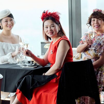 Women dressed in race day fashion and fascinators celebrating with a toast at a table at Eureka 89, seated against floor to ceiling windows. 
