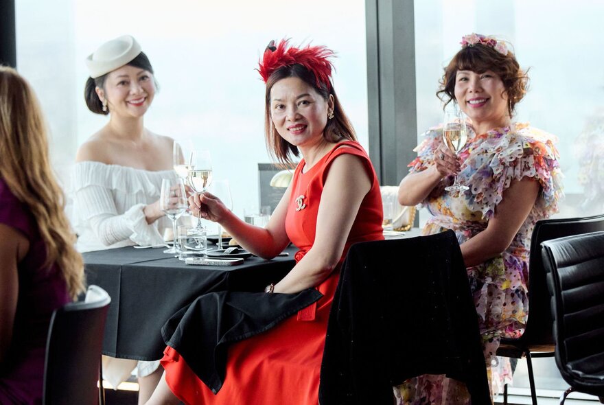Women dressed in race day fashion and fascinators celebrating with a toast at a table at Eureka 89, seated against floor to ceiling windows. 