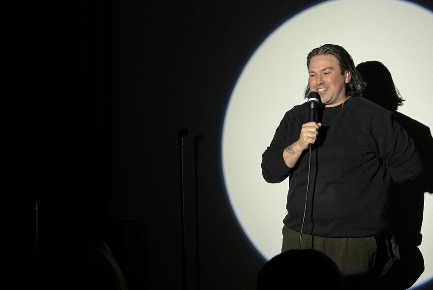 A stand up comedian on a stage, holding a microphone, performing in a spotlight.