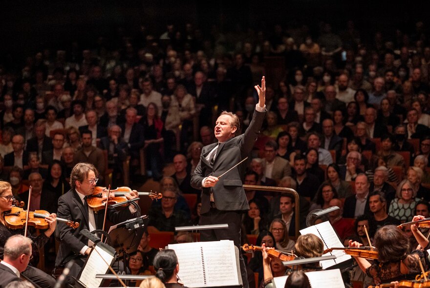 An orchestra on a stage, playing their instruments, with a conductor in front of them and a seated audience in the background.