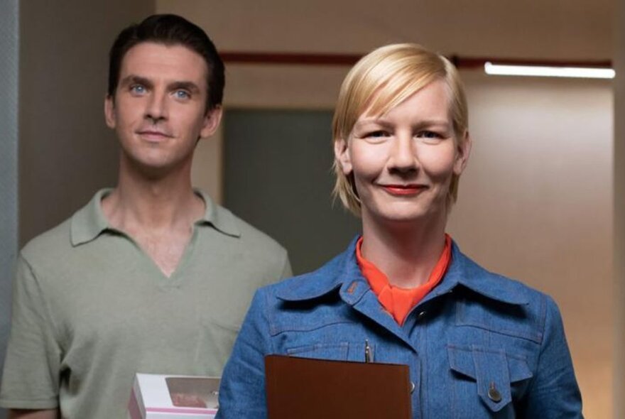 A man and woman looking ahead, the woman wearing a military-style shirt and carrying a folder, the man standing behind looking quizzical.