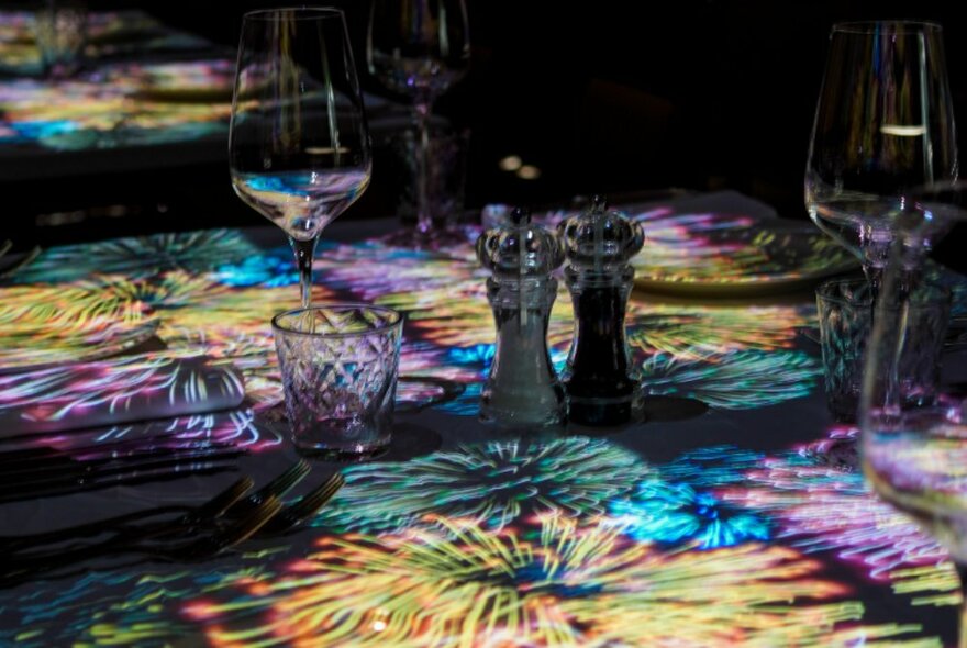 Dining table set for dinner with glasses and condiments on a fireworks-themed tablecloth.