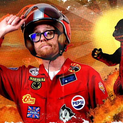 A man in a red racing suit with patches sewn all over it, wearing a helmet and glasses with a cartoon godzilla in the background.